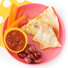 A pink kids plate with pizza quesadillas cut grapes and peppers with a white and yellow napkin against a white background.
