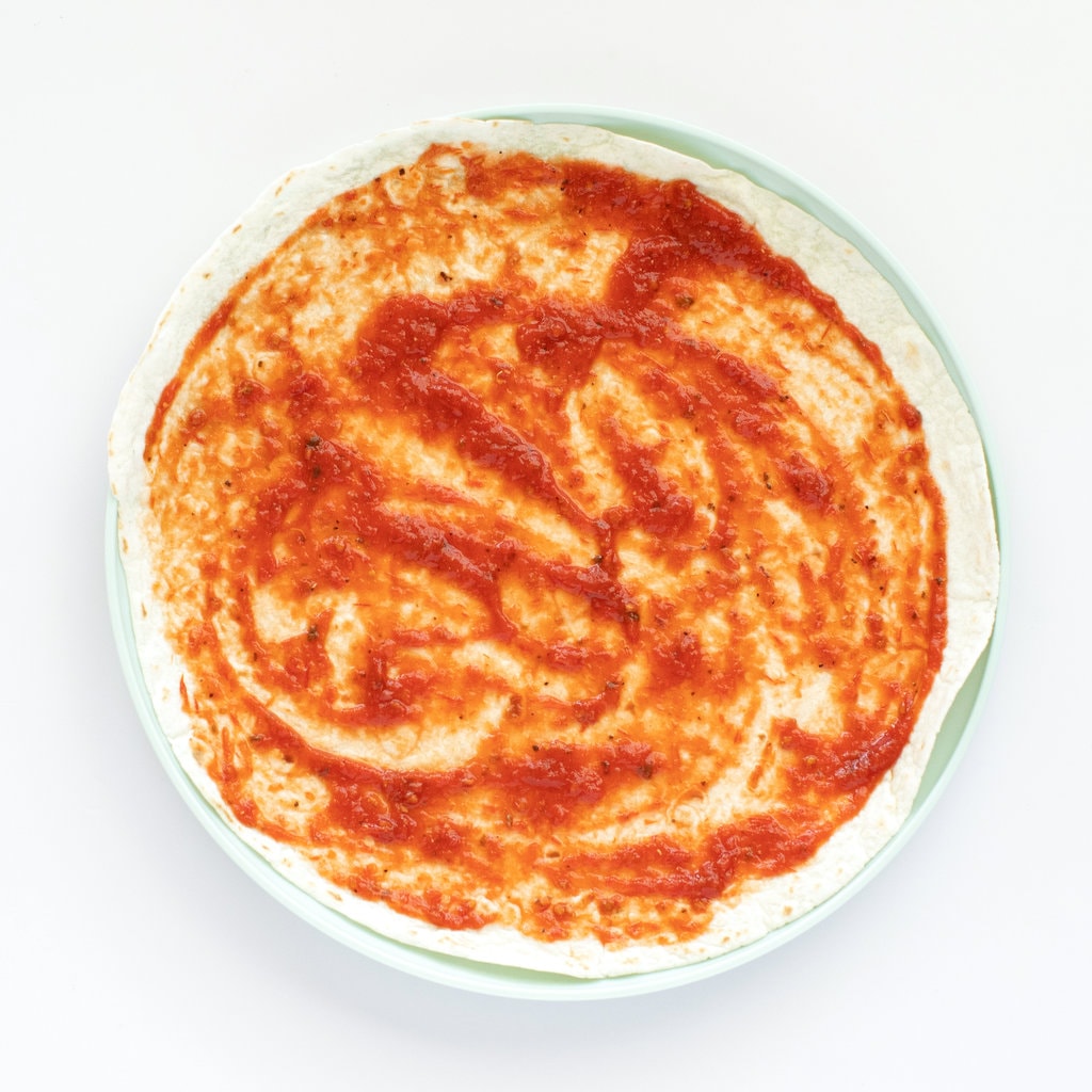 A tortilla with pizza sauce on top sitting on a teal plate.