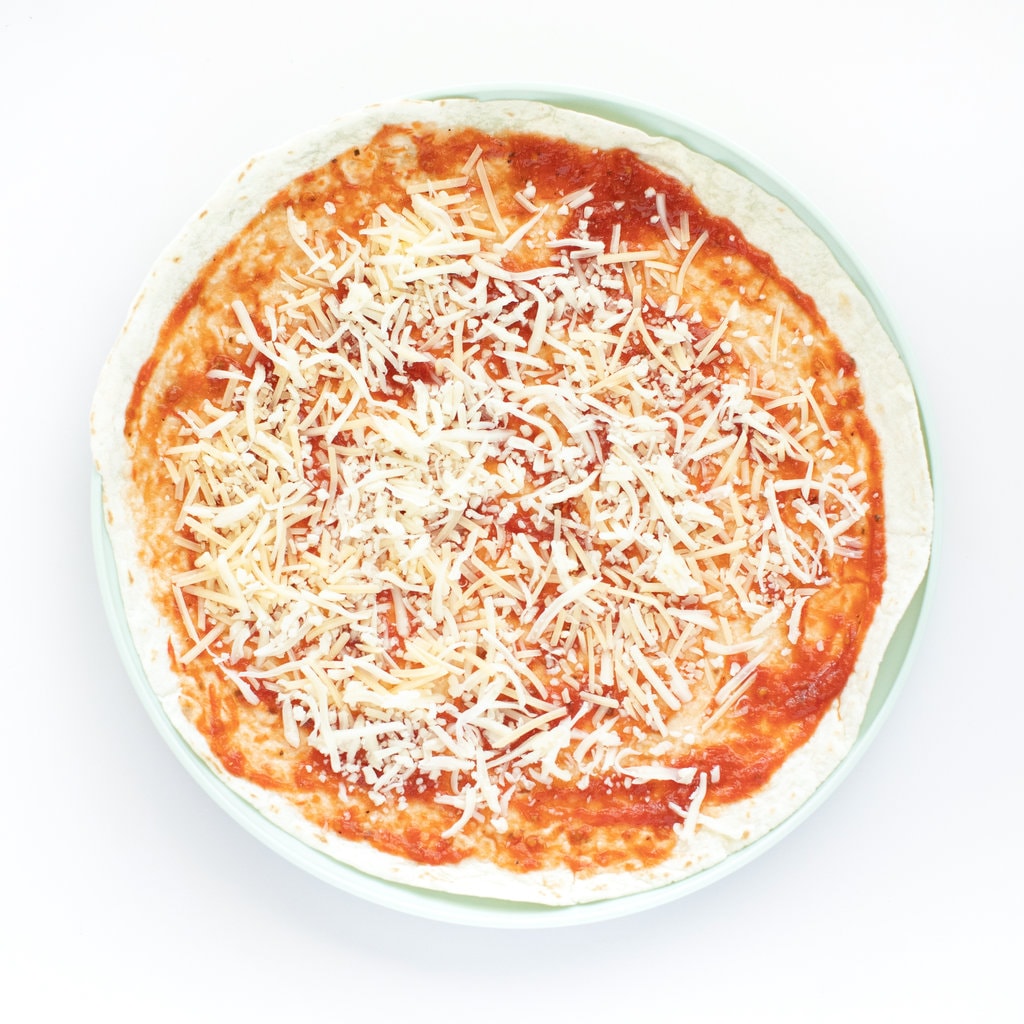 A tortilla with pizza sauce and cheese sitting on a two plate with a white background.