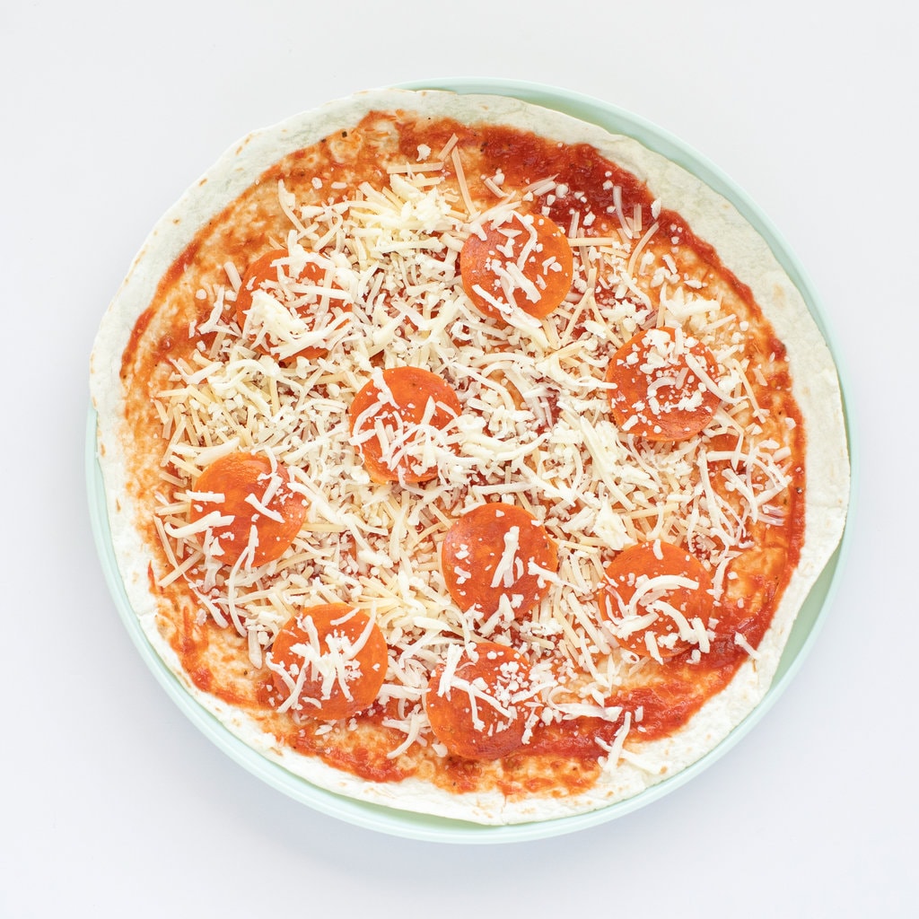 A tortilla with pizza sauce cheese and pepperoni on a teal plate with a white background.