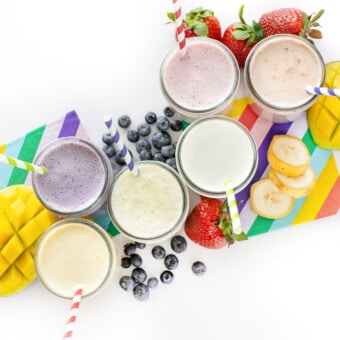 An overhead shot of different glass cups filled with flavored milk with fruit scattered around and colorful napkins against a white background.