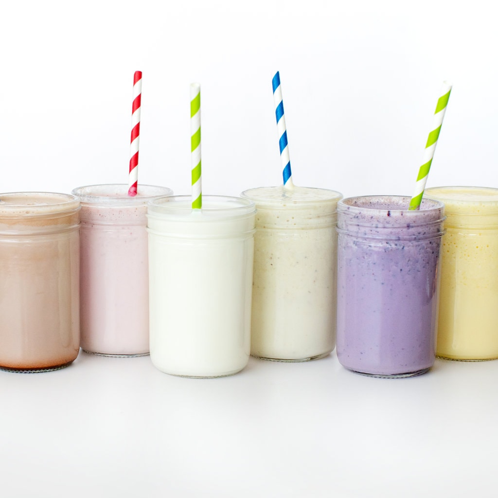 A line of clear glass cups filled with different flavors of milk – chocolate milk, strawberry milk, vanilla milk, mango milk, blueberry milk, and mango milk with colorful straws coming out against a white background.