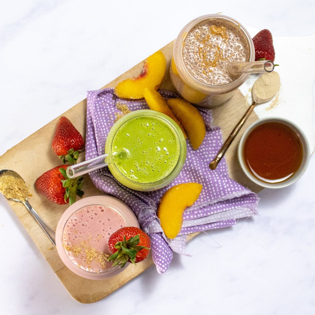 A wooden cutting board with three different flavors of a lactation smoothie, one is strawberry, one is spinach peach, one is chocolate peanut butter all sitting on a marble countertop.