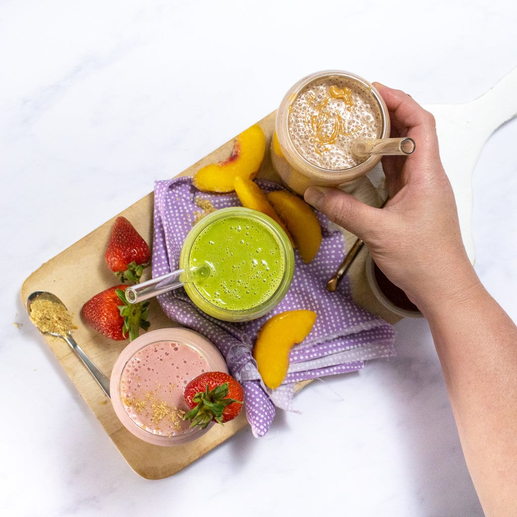 A wooden cutting board with three different flavors of a lactation smoothie, one is strawberry, one is spinach peach, one is chocolate peanut butter all sitting on a marble countertop.