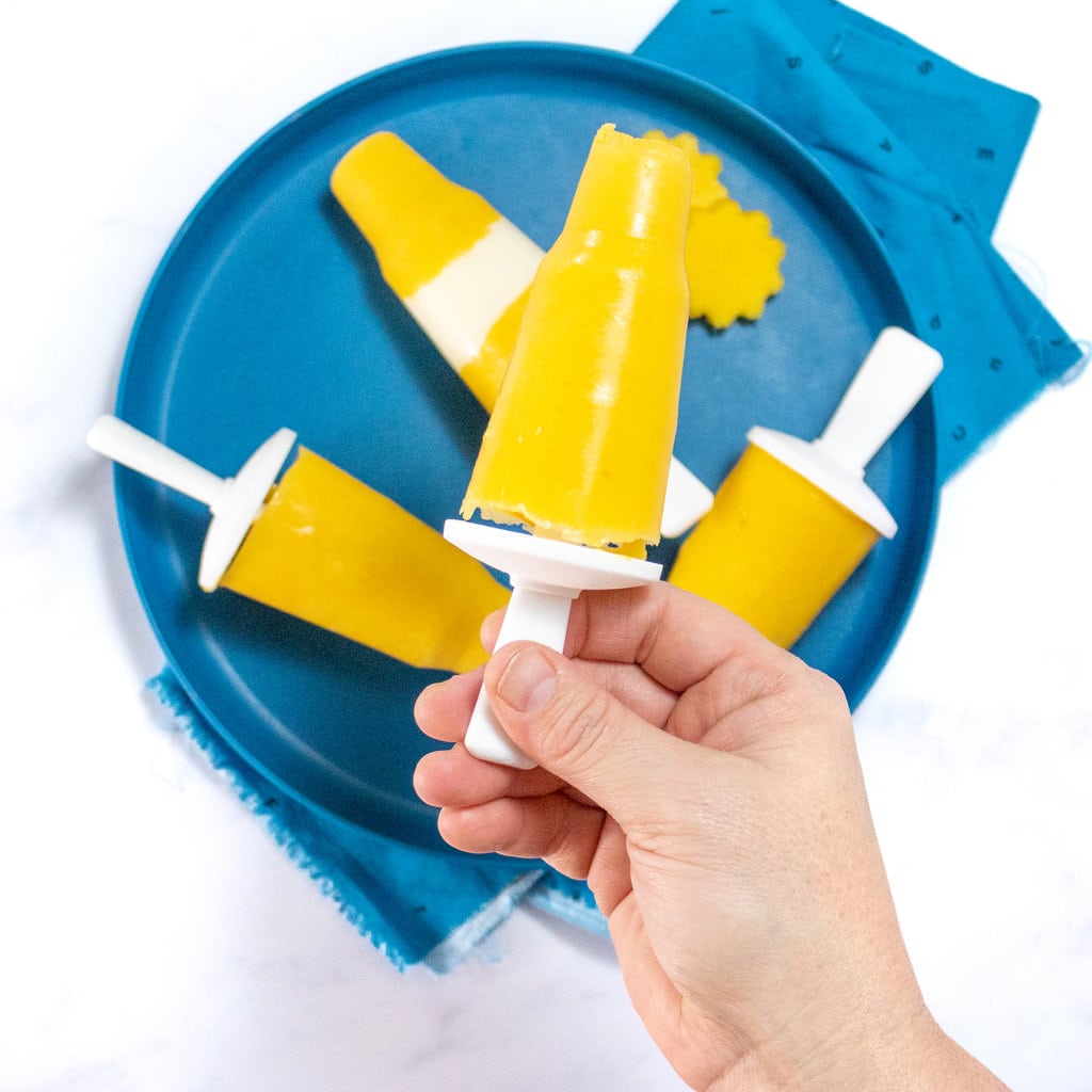 A hand holding a mango popsicle over a teal plate full of mango popsicles with yogurt.