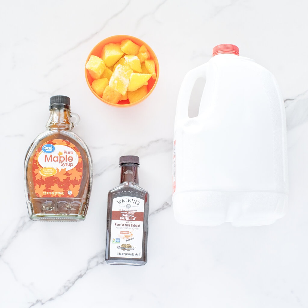 Ingredients for mango milk countertop – milk, vanilla extract, maple syrup and mangoes. Against a white marble background.