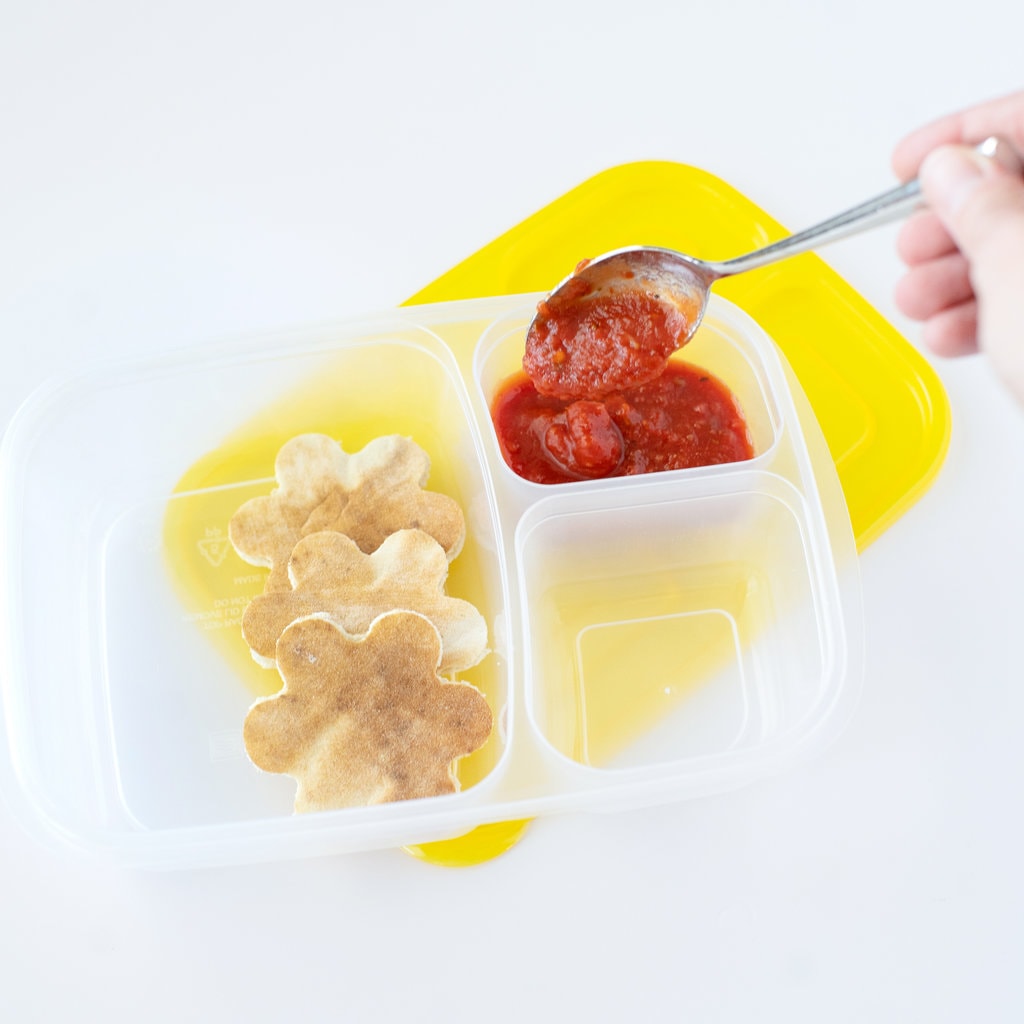 A woman's hand spooning in pizza sauce into a container in a lunchbox for a kids lunchable.