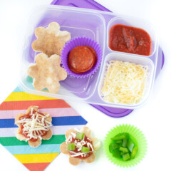 A kids lunch container packs with pizza crust, shredded cheese, and tomato sauce to make homemade pizza for lunchables, along with a colorful striped napkin.