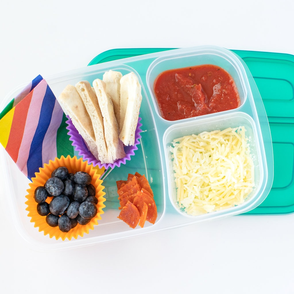 A kids lunchbox with a colorful lid with containers full of shredded cheese, pizza sauce, strips of pita bread, blueberries and chopped pepperoni with a colorful napkin against a white background.
