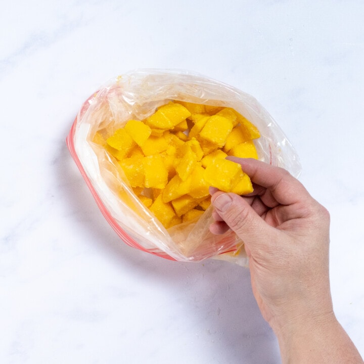 A clear Ziploc bag with frozen mango chunks with a hand holding up a chunk over a white marble countertop.