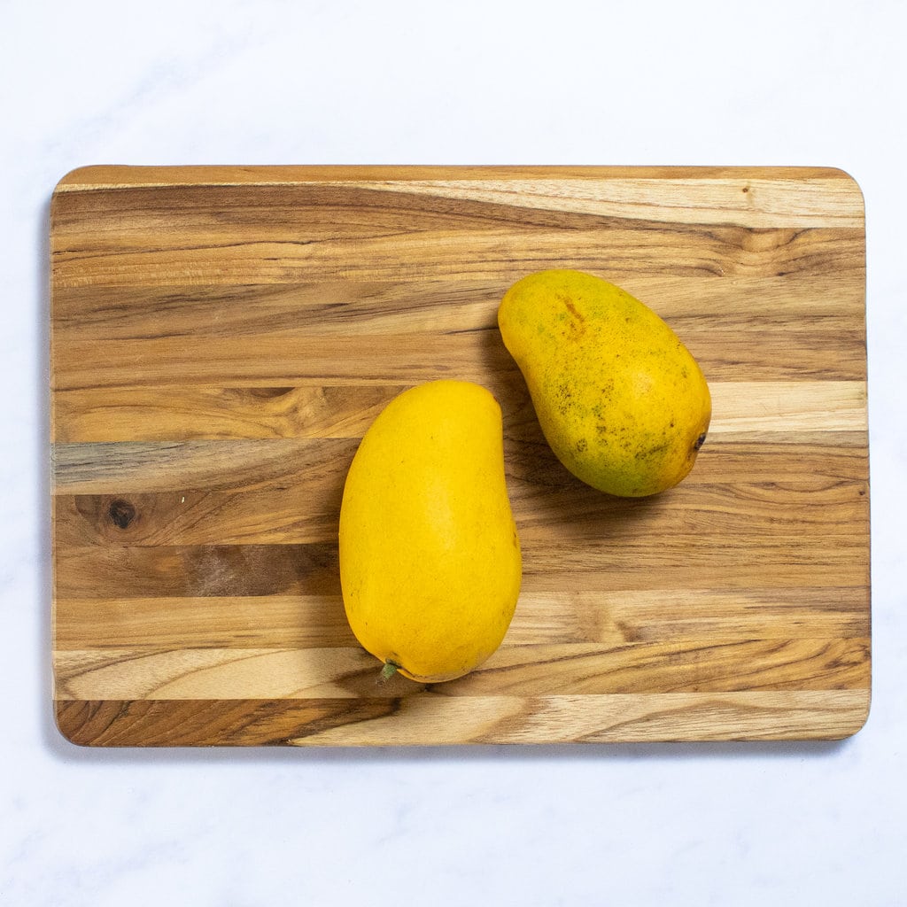 Two mangoes on the wooden cutting board sitting on a white marble counterto