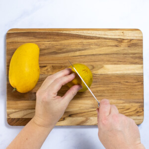 Someone cutting a mango of the pit on a wooden cutting board. Someone Someone
