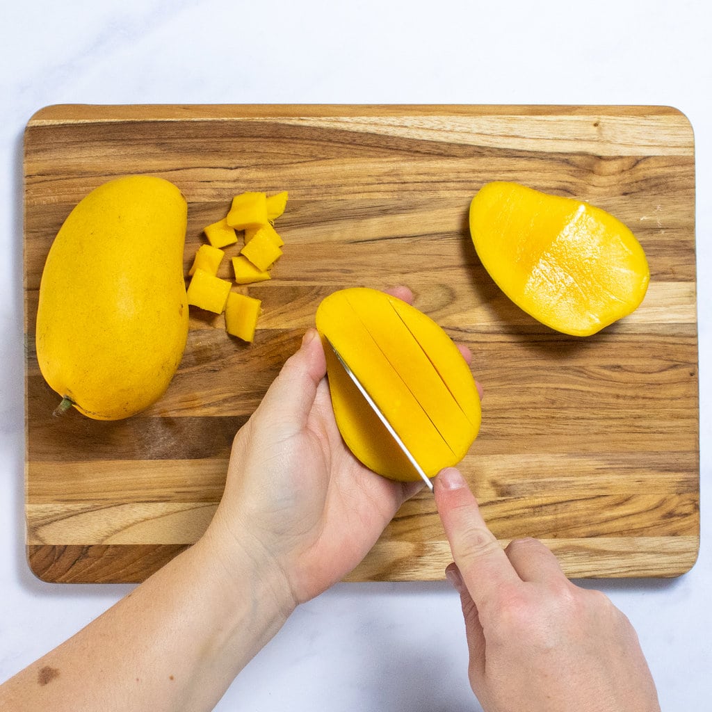 Two hand slicing a mango into chunks over a wooden cutting board.