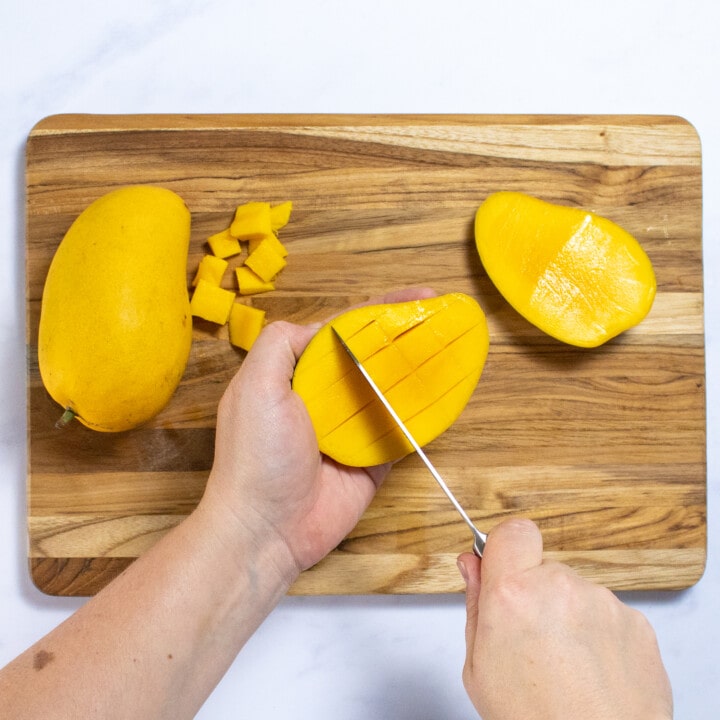 Two hand slicing a mango into chunks over a wooden cutting board.