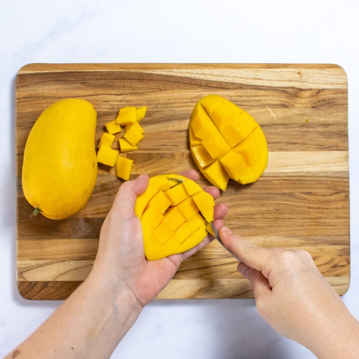 A wooden cutting board with two hands cutting up a mango.