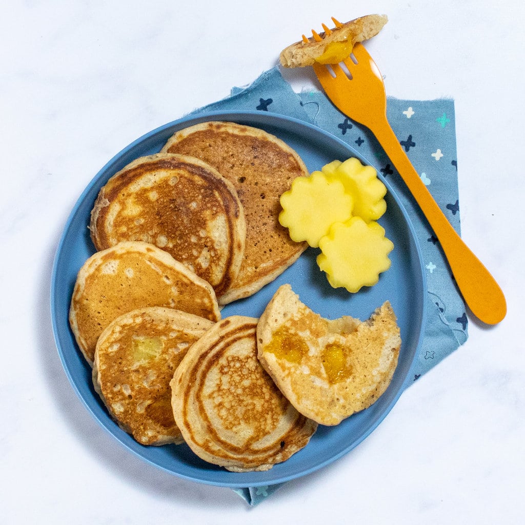 A blue kids plate with three mango pancakes and mangoes cut into shapes with a blue colorful napkin and an orange fork on a white background.