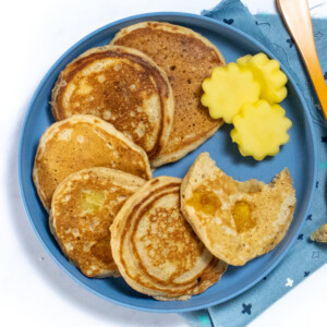 A blue kids plate with three mango pancakes and mangoes cut into shapes with a blue colorful napkin and an orange fork on a white background.