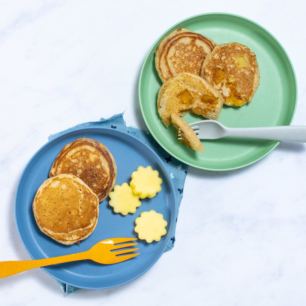 Two blue and green kids plates with mango pancakes and kid forks that are colorful on a marble background.