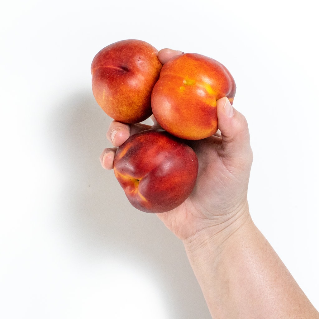 Hands holding three nectarines against away background.