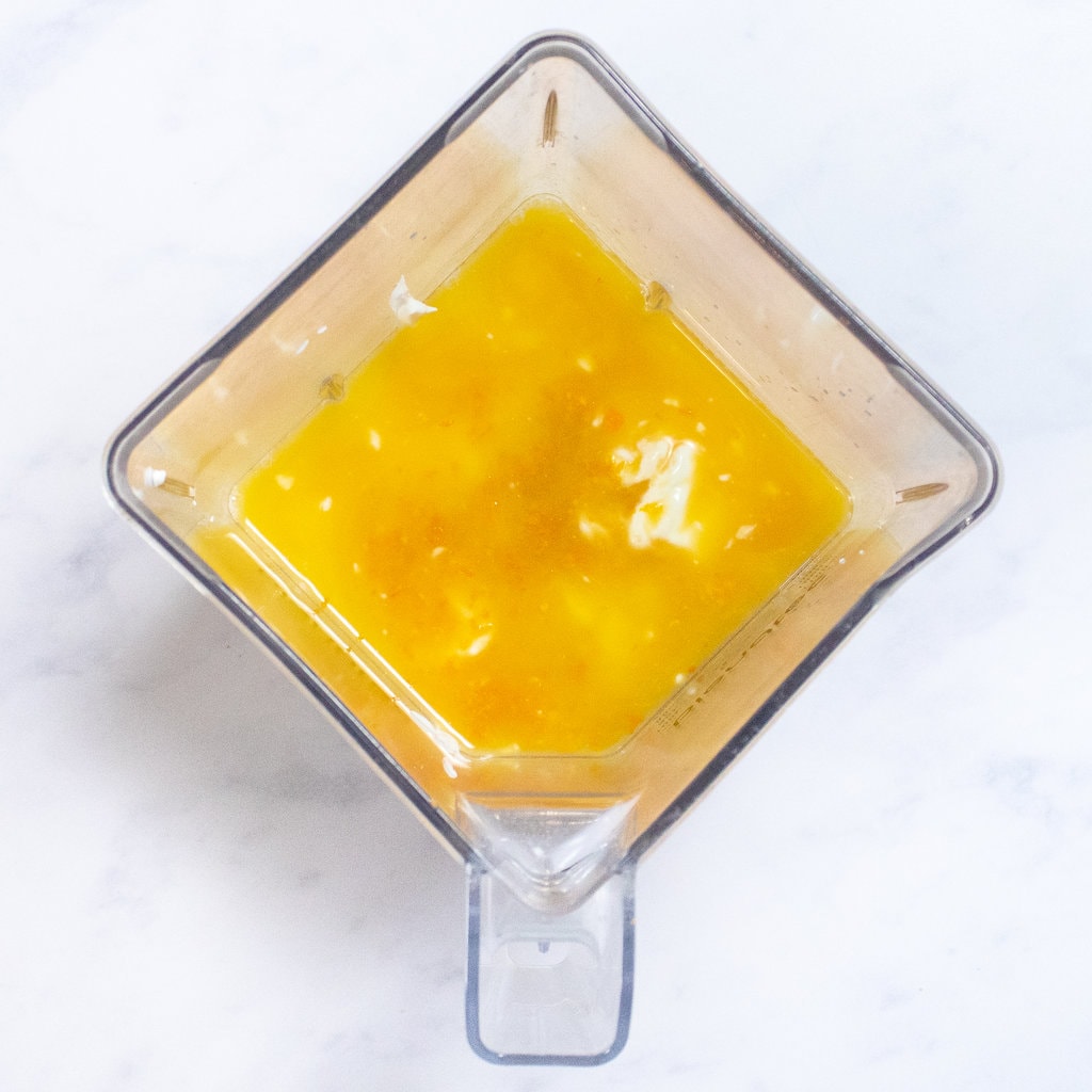 A clear blender on a white marble countertop full of ingredients for orange Popsicles.