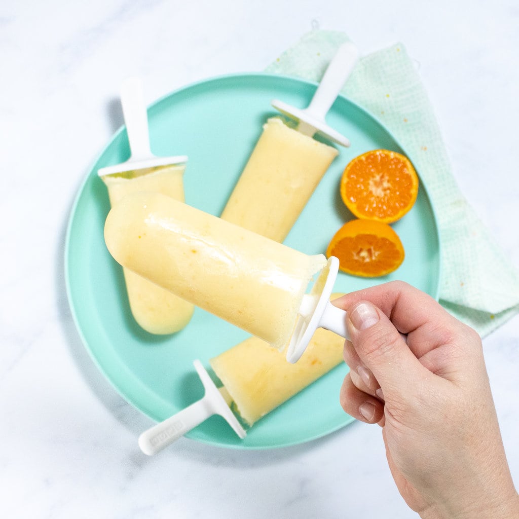 A hand holding an orange Creamsicle popsicle over a teal plate with orange slices on a white marble countertop.