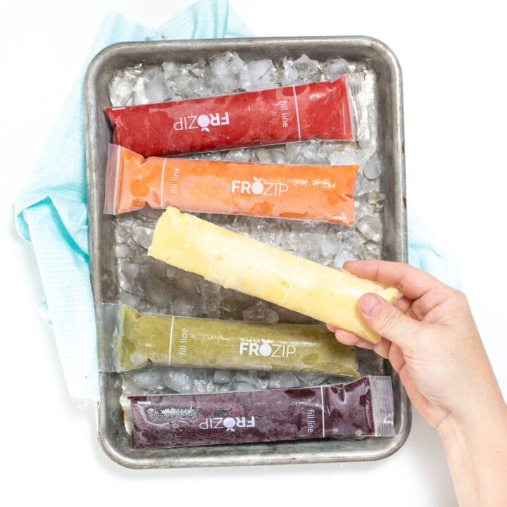 A hand holding a yellow freezer, pop against a baking sheet of ice along with other rainbow colored freezer pops.