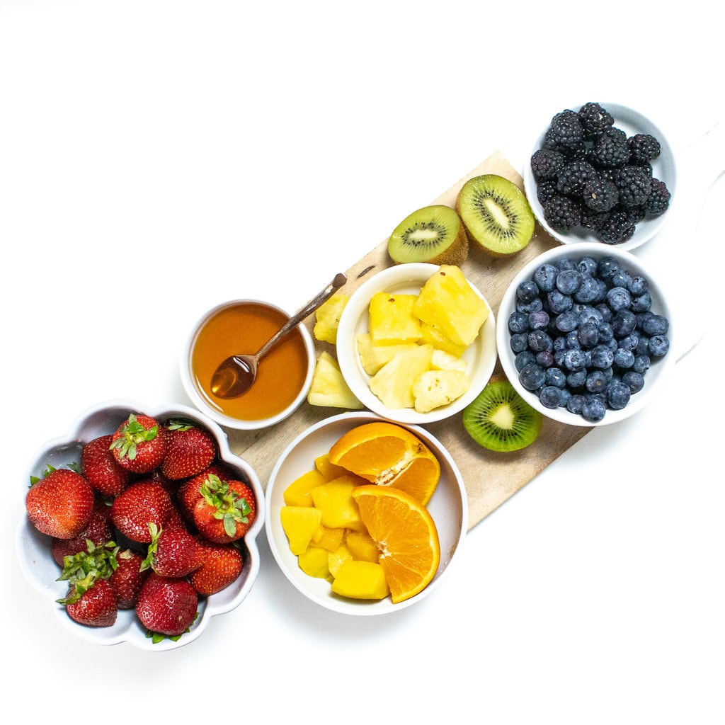 A spread of a fruit and a rainbow color with a small bowl of honey I guess the way background.