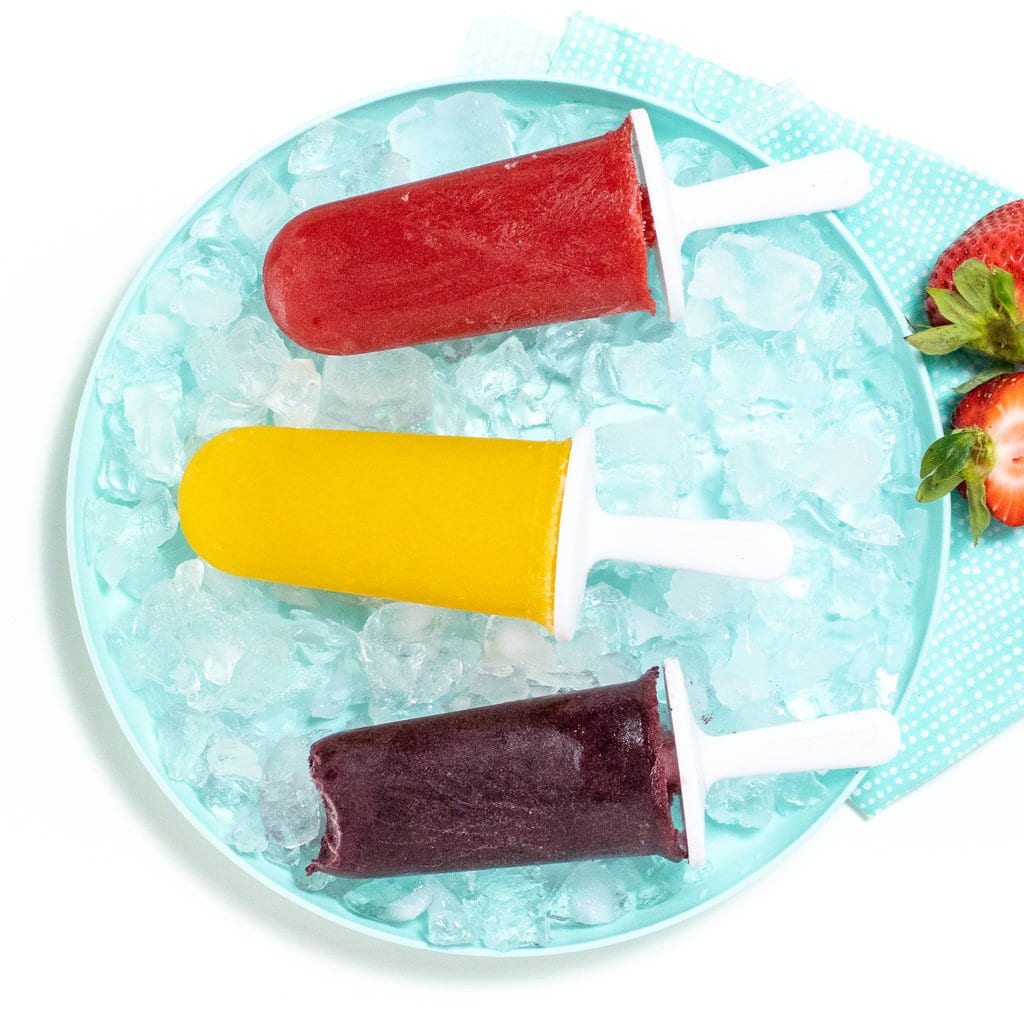 strawberry popsicles, mango popsicles and a grape popsicle all lined up on a kids blue plate with fruit off to the side.