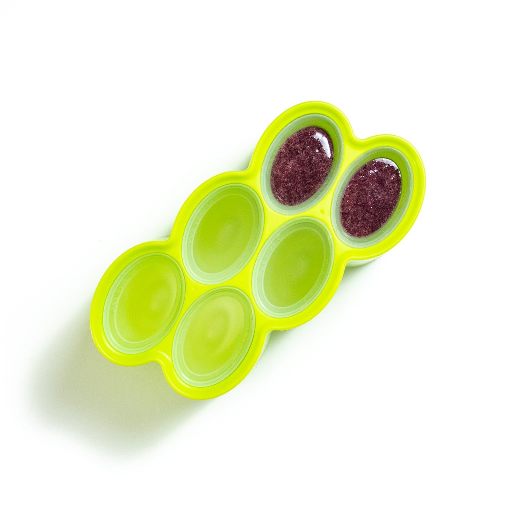 A green popsicle mold with 2 molds full of a dark purple fruit mixture.