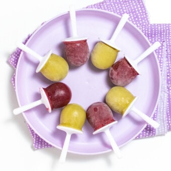 A purple kids plate full of alternating purple and green grape Popsicles in a purple napkin against a white background.