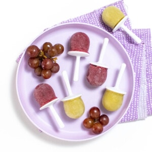 A purple kids plate full of both purple and green grape Popsicles with grapes on the plate against a white background.