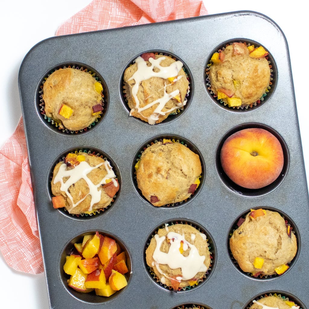 A muffin tray filled with peach muffins, a peach, and some chopped peaches, some of the muffins have a cream cheese drizzle against a white background.