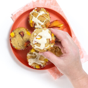 A hand holding a peach muffin with a cream cheese drizzle over a pink kids plate with a couple of peach muffins on top.