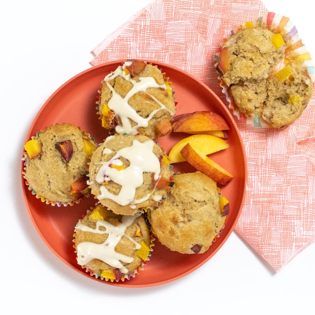 A pink kids playing with a pink napkin with peach muffins on top slices of peach and a muffin cut in half to show the fluffy interior.