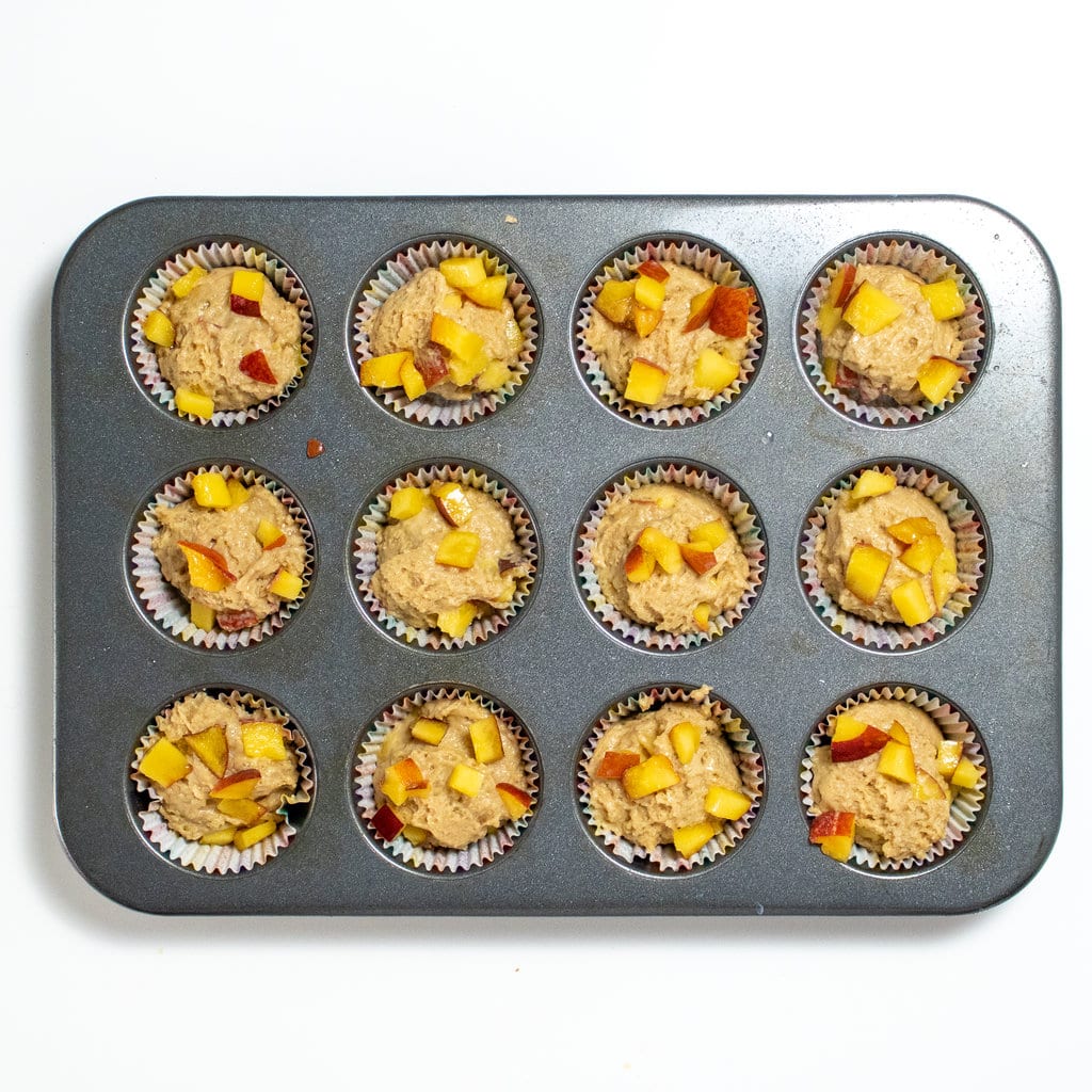 A silver muffin tin against a white background bowl with cooked peach muffins with peaches on top.
