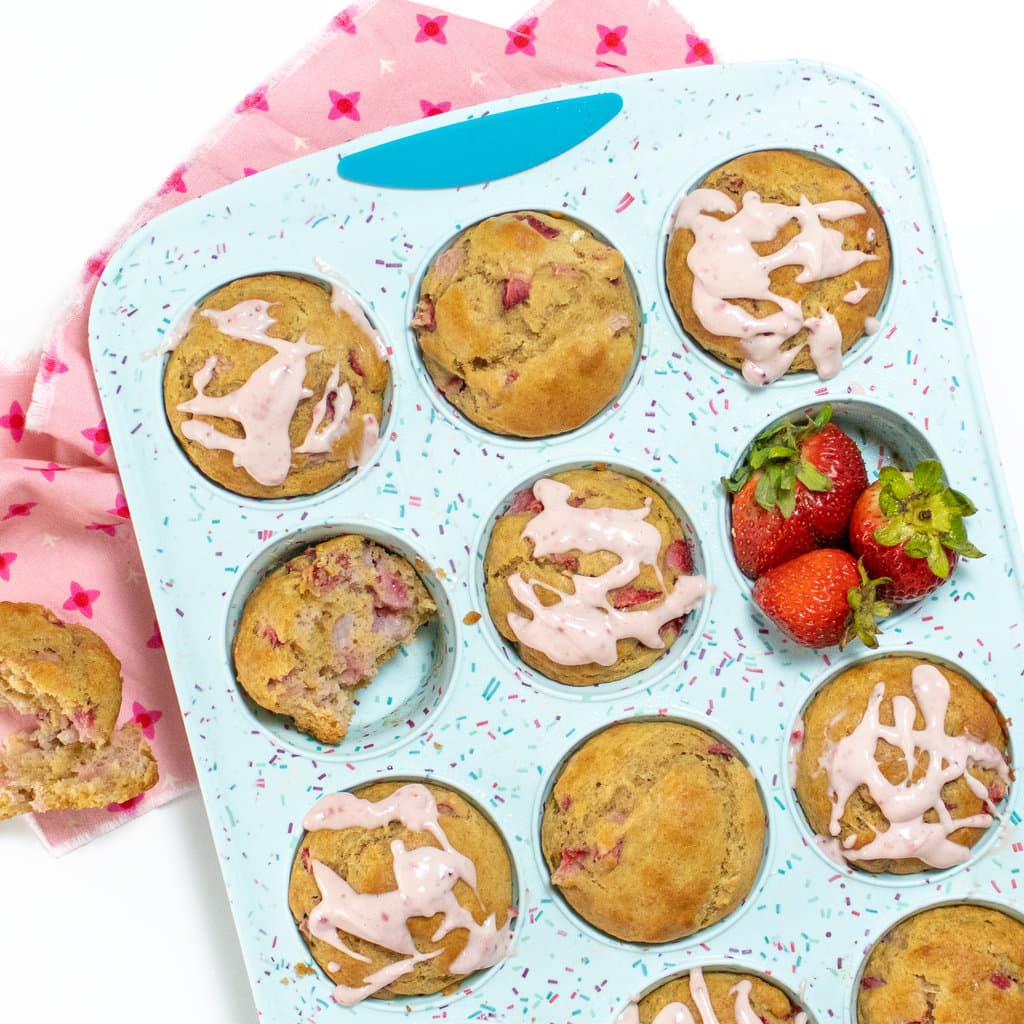 A blue muffin tin full of strawberry muffins with a cream cheese, strawberry, drizzle with a side of strawberries, a pink napkin, and a white background.