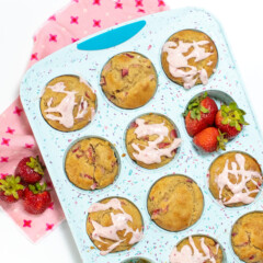 A blue muffin tin full of strawberry muffins with a cream cheese, strawberry, drizzle with a side of strawberries, a pink napkin, and a white background.