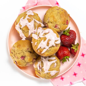 A kids, pink plate full of healthy strawberry muffins, some with a strawberry cream cheese, drizzle, and a side of strawberries, with a pink napkin on a white countertop.