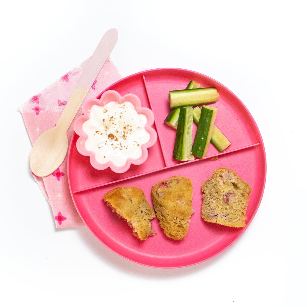 A pink kids play with three sections, one is with a strawberry muffin cut into thirds, one of them cut cucumbers, and another is of a yogurt in it with a sprinkle of cinnamon on top. On the side is a pink wooden spoon and a pink napkin.