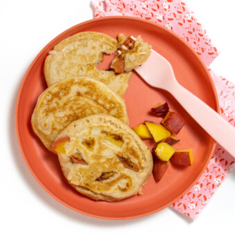 A pink kids plate with three peach pancakes and some chopped up peaches with a pink fork against a white background.