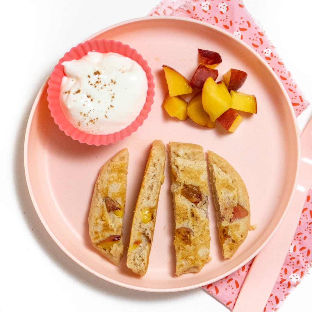 Play pink kids play with cut up strips of peach pancakes, chunks of peaches, and a pink bowl of yogurt.