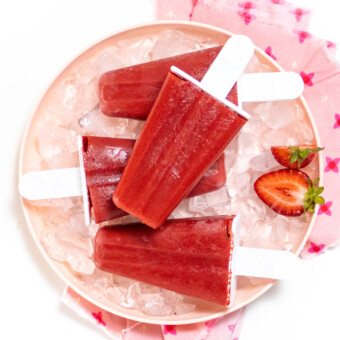 A kids pink white background, the plate has four strawberry popsicle stack on top of each other and strawberries cut on the side.