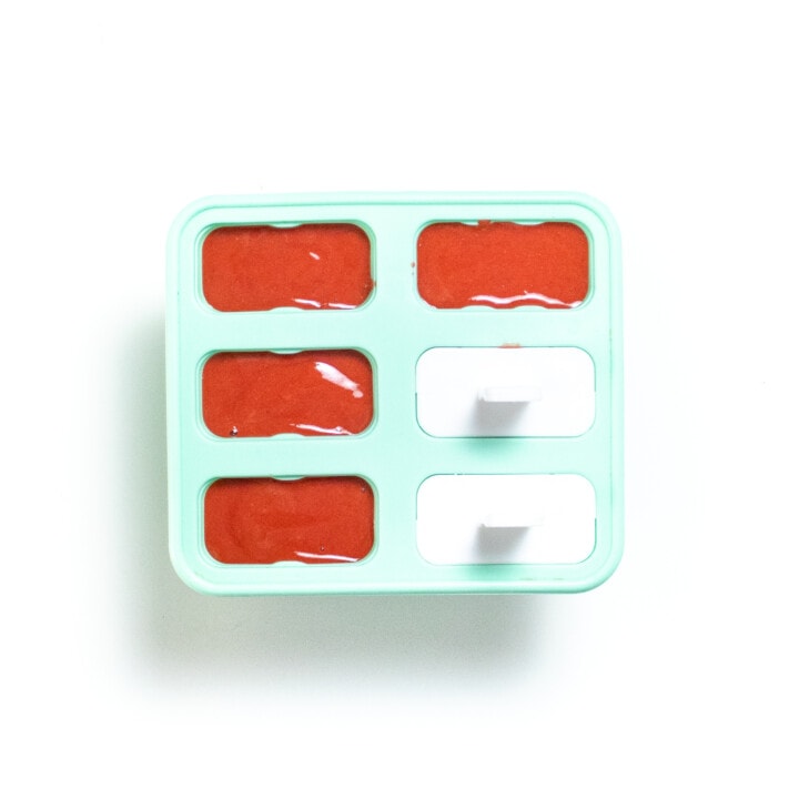 A teal popsicle tray against a white background full of strawberry purée.