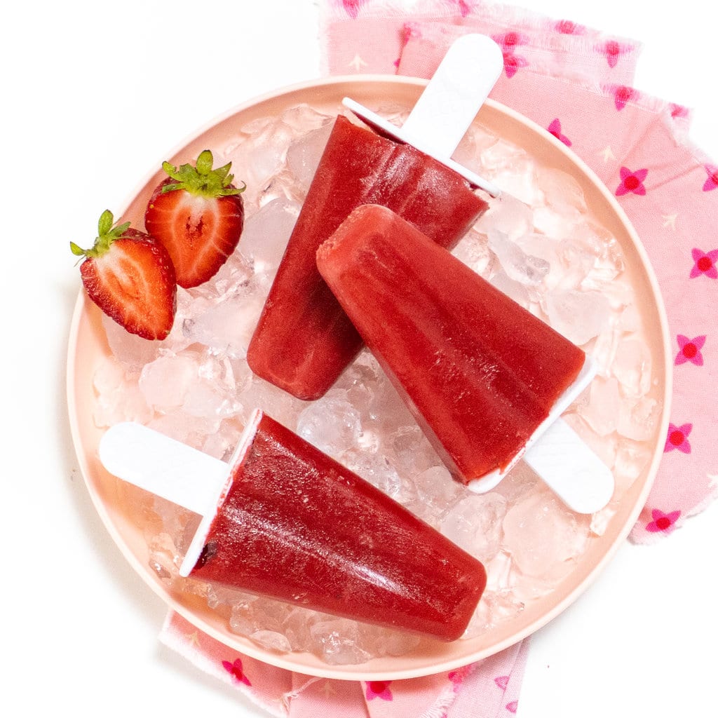 A pink kids plate with ice cubes and three strawberry popsicles, with strawberries on the side in a pink napkin against a white background.