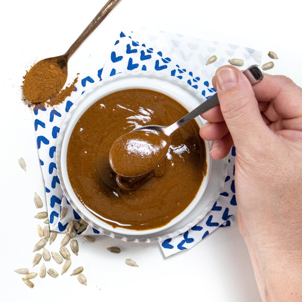 A white bowl of sun butter, with a hand, holding a spoon pulling some out.