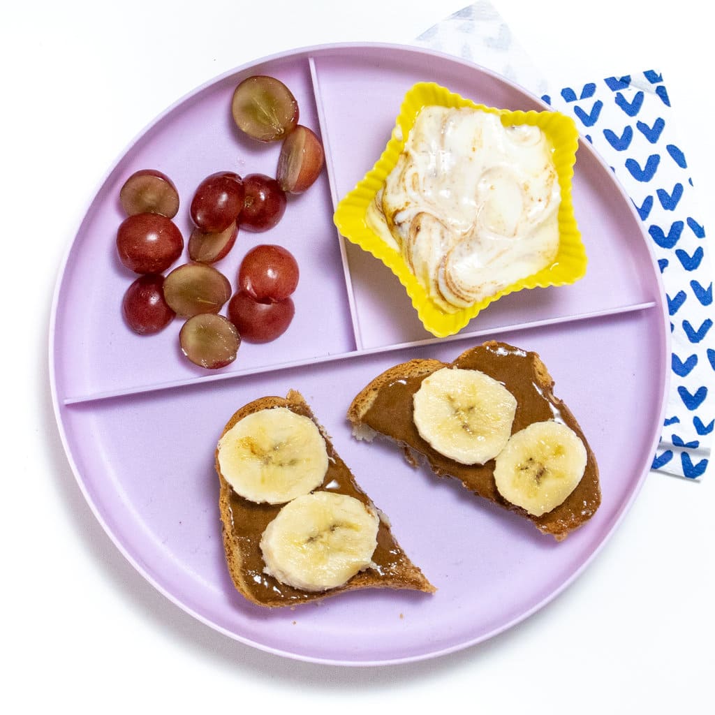 A kids, purple plate, full of sun, butter, toast, with banana, a side of yogurt, with cinnamon, and cut grapes against a white countertop.