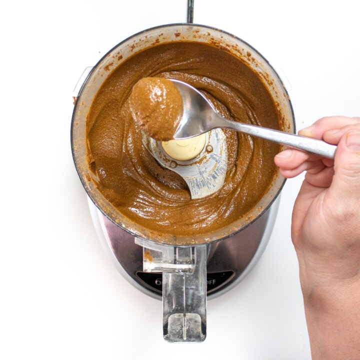 A food processor with a hand, holding a spoon over full of not quite done sun butter.