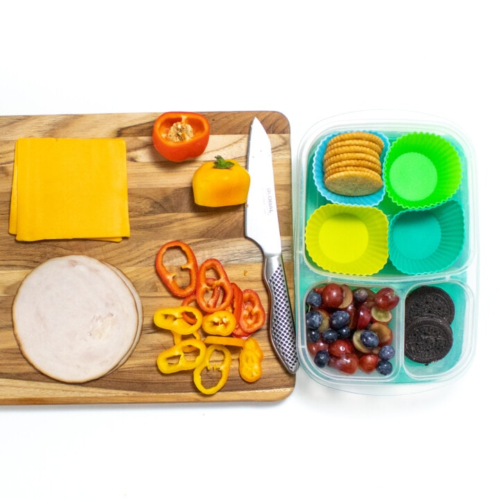 A wooden cutting board on a white countertop with fruits and veggies cut and ready to be put into a kids lunchbox.