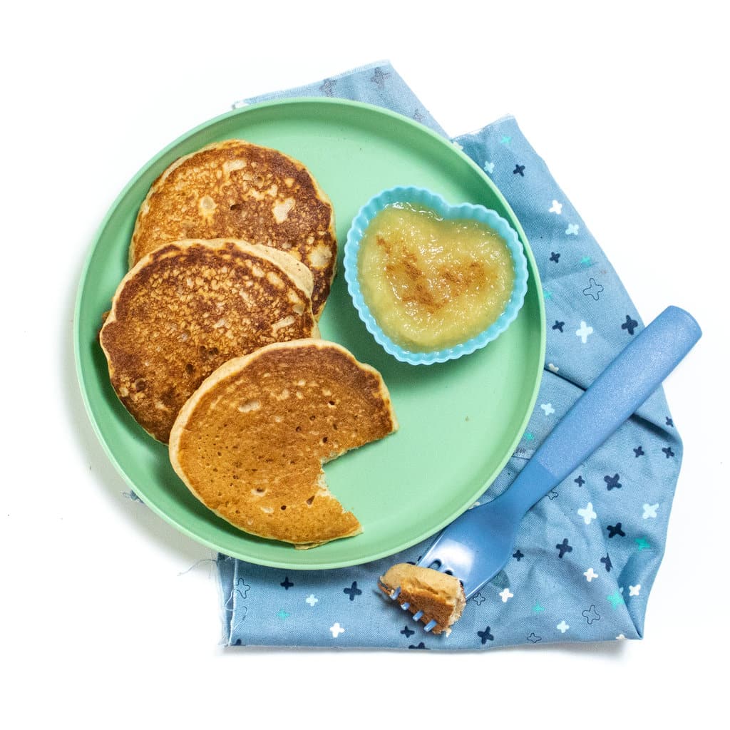Teal kids plate with 3 applesauce pancakes, a small heart shaped bowl for applesauce, a blue fork and a blue patterened napkin.