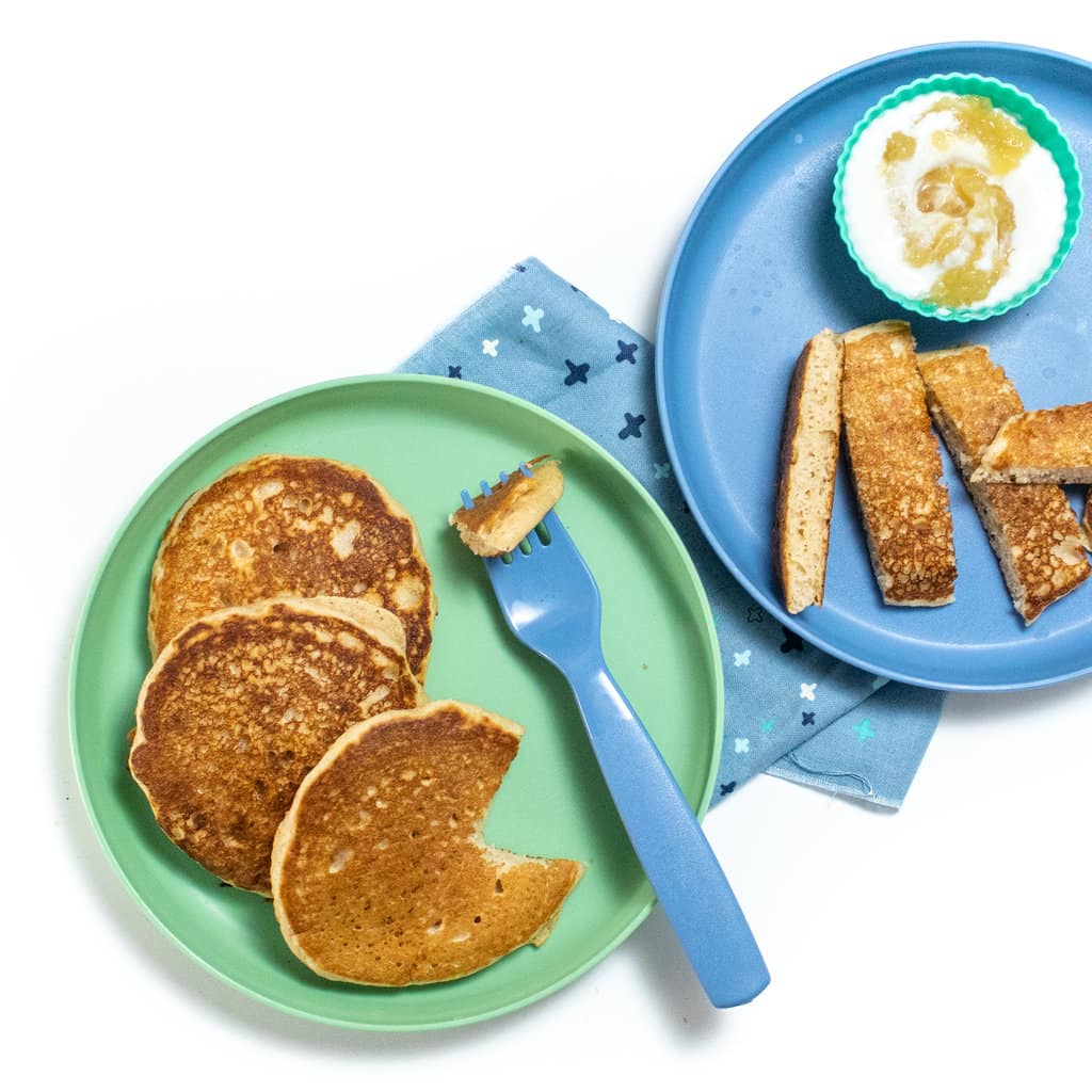 2 blue kids plates with applesauce pancakes for kids, cut into different shapes with a small bowl of yogurt and applesauce and a fork and blue napkin.
